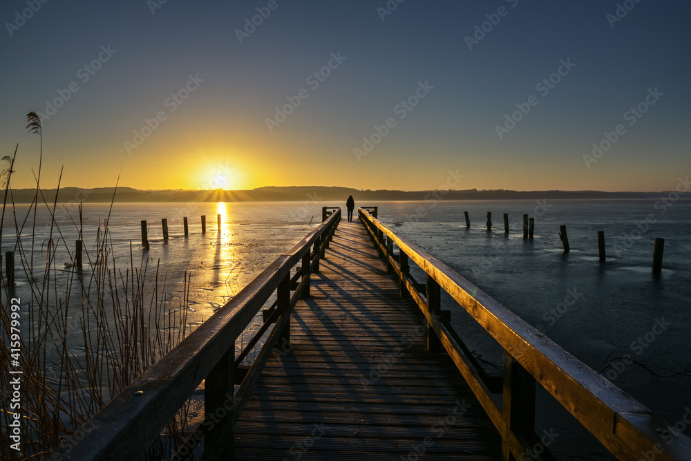 Sunrise over a frozen lake and a young woman as silhouette on the end of a long wooden jetty enjoying the landscape scenery, copy space,