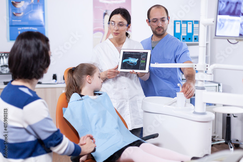 Dentist in dentistiry office showing tablet pc with jaw radiography to parent and little girl explaining treatment. Stomatologist explaining teeth diagnosis to mother of child in health clinic holding
