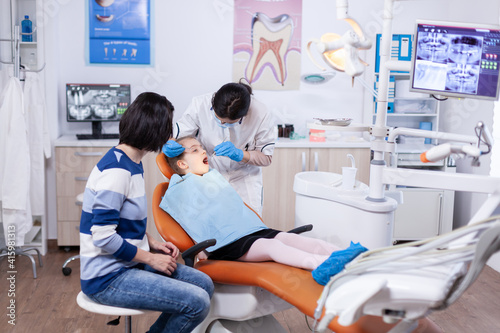 Kid patient in dentistiry office getting treatment for teeth cavity sitting on dental chair wearing bib. Dentistry specialist during child cavity consultation in stomatology office using modern