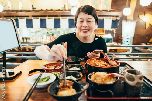 Wall Mural Young asian korean woman eating side dishes at japanese local restaurant and texting on smartphone