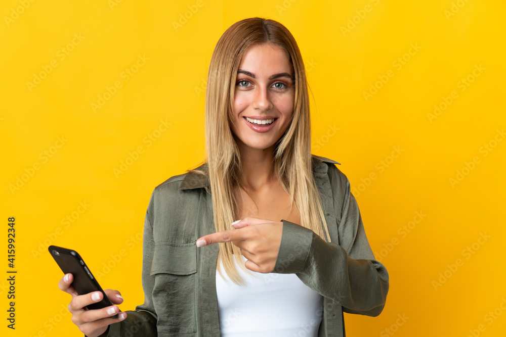 Young blonde woman isolated on yellow background using mobile phone and pointing it Young blonde woman isolated on yellow background using mobile phone and pointing it