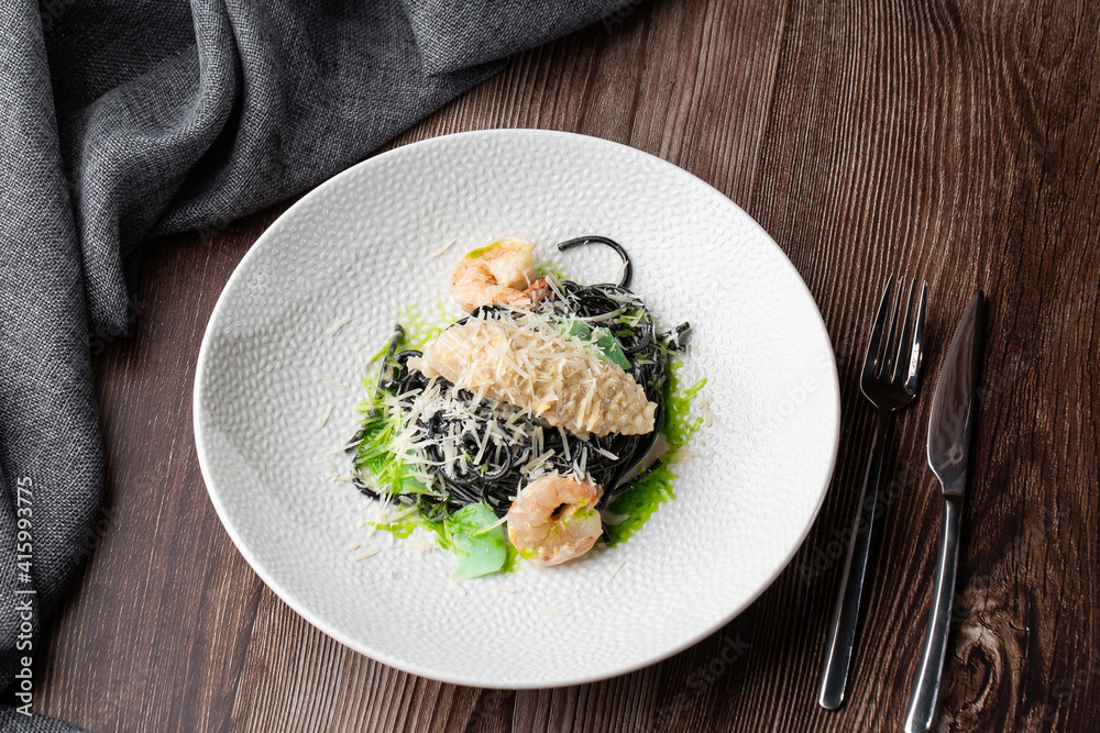 Black spaghetti. Black seafood pasta with shrimps and parmesan on white plate isolated on wooden background. Silver cutlery and grey linen napkin by side. Mediterranean gourmet food. Black spaghetti. Black seafood pasta with shrimps and parmesan on white plate isolated on wooden background. Silver cutlery and grey linen napkin by side. Mediterranean gourmet food.