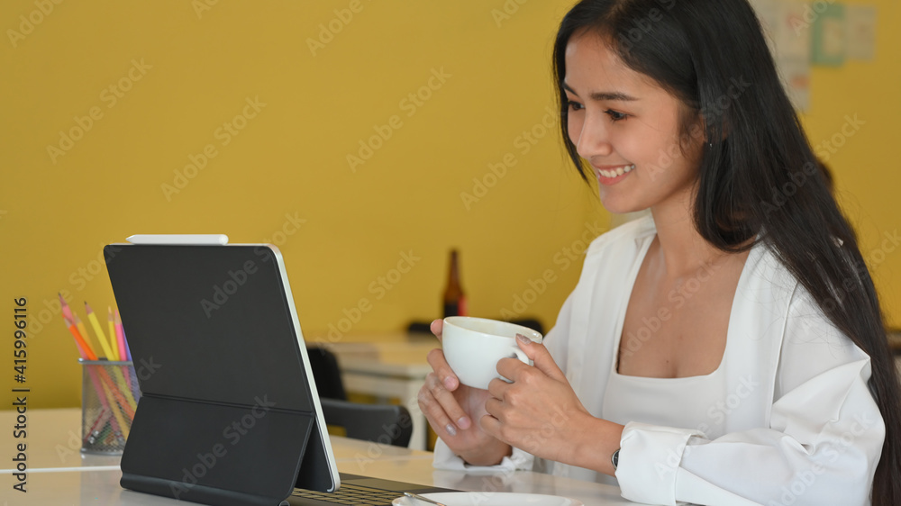 Young beautiful female holding coffee cup and smiling while looking on digital tablet Young beautiful female holding coffee cup and smiling while looking on digital tablet