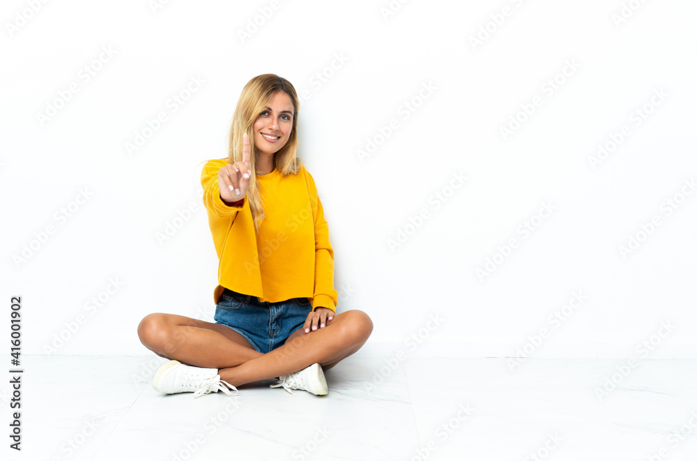 Young blonde Uruguayan woman sitting on the floor isolated on white background showing and lifting a finger Young blonde Uruguayan woman sitting on the floor isolated on white background showing and lifting a finger
