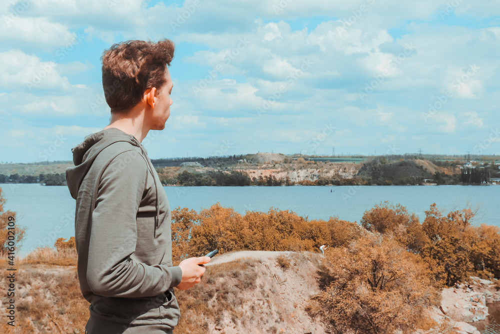 A young guy in a sweatshirt holds a phone in his hands and enjoys the view of the quarry and the lake on a warm spring day.