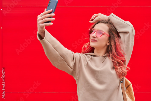 Young blogger woman with pink dyed hairs staying near the red wall and making selfie by smartphone