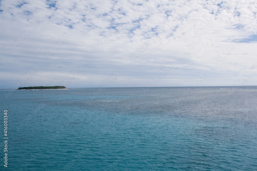 Lady Musgrave Island, a remote Great Barrier Reef atoll far offshore. Blue ocean with shadows of the underwater coral reef and the coral island on the horizon under a sky of white clouds.