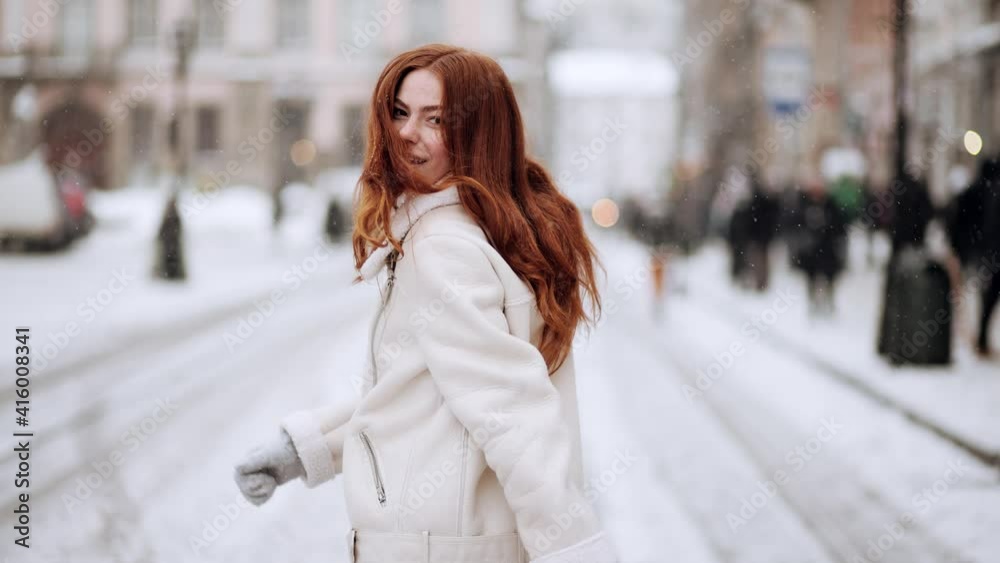 Young smiling red hair woman with lovely freckles run and spinning in the city street. Attractive fashion model with flying hair. Beautiful girl shakes hair. Snowing outdoors. Winter concept Young smiling red hair woman with lovely freckles run and spinning in the city street. Attractive fashion model with flying hair. Beautiful girl shakes hair. Snowing outdoors. Winter concept