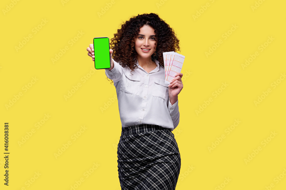 Excited young curly girl holding lottery tickets and smartphone with blank green screen isolated over yellow background Excited young curly girl holding lottery tickets and smartphone with blank green screen isolated over yellow background