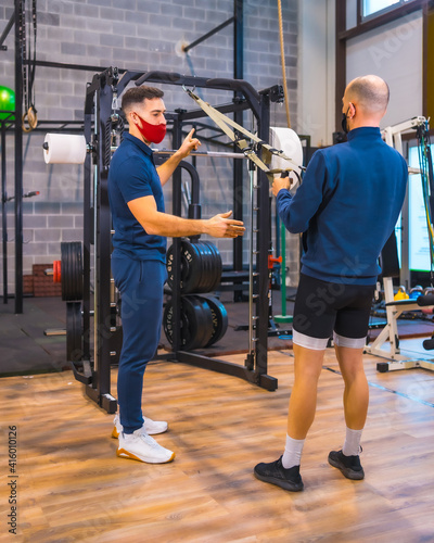 A young athlete with an instructor in the gym doing arm exercises in the coronavirus pandemic, a new normal. With protective face mask