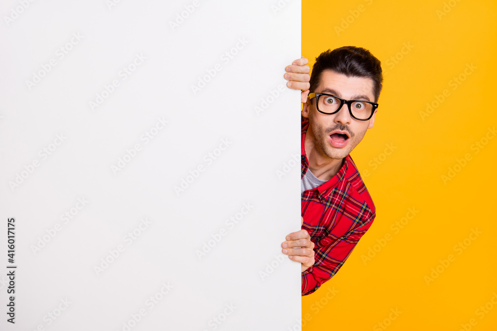 Photo of amazed shocked man hold hide behind white board copyspace isolated on yellow color background