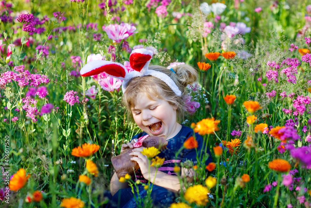 Portriat of adorable, charming toddler girl with Easter bunny ears eating chocolate bunny figure in flowers meadow. Smiling happy baby child on sunny day with colorful flowers, outdoors.