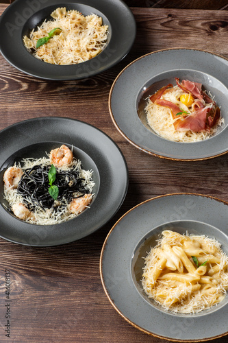 Four kinds of different cooked pastas: Penne, Farfalle, Carbonara, Black Spaghetti. Close-up isolated on wooden background in black and grey plates. Mediterranean gourmet food. Top view. © MONIUK ANDRII