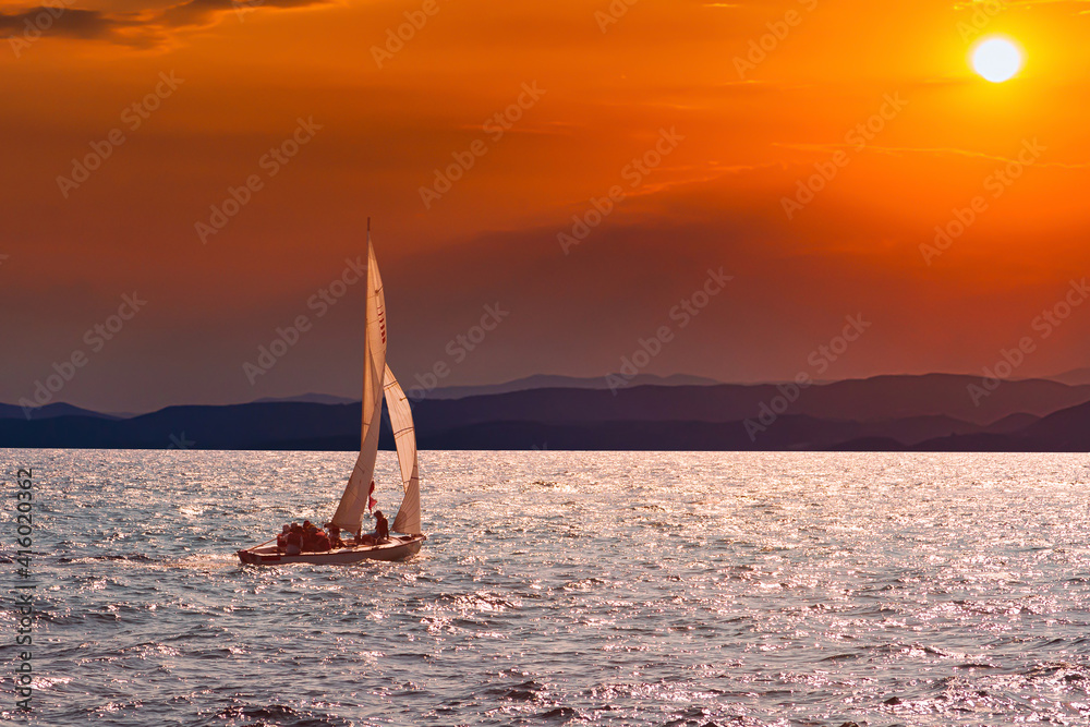 Sailboat sails over the sea in on the background of a beautiful sunset red sun. Sea on the horizon rests on the mountains. Beautiful sunset on the background of mountains