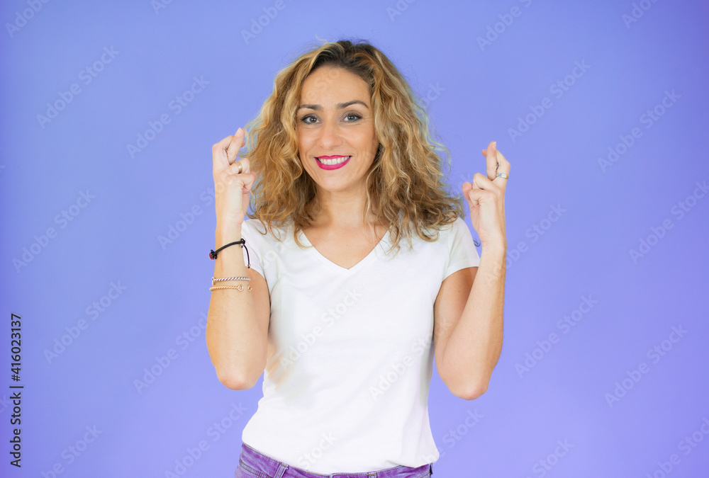 Beautiful woman with curly hair wearing white casual t-shirt over isolated background gesturing finger crossed smiling with hope. Luck and superstitious concept.