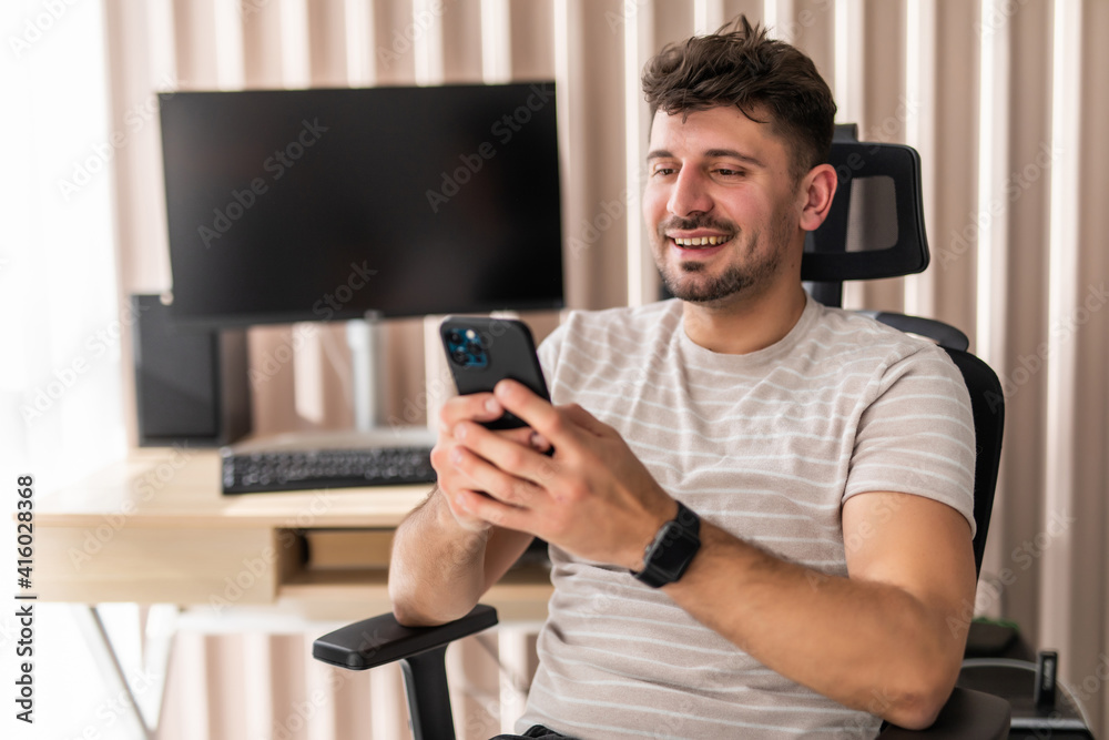 Handsome young man looking at his smart phone and smiling while sitting in big comfortable chair at home Handsome young man looking at his smart phone and smiling while sitting in big comfortable chair at home