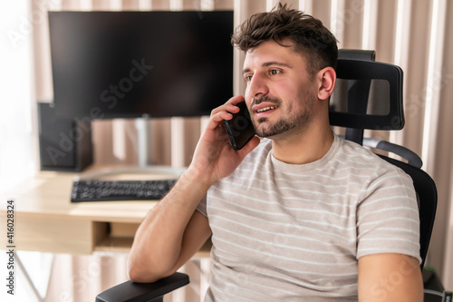 Working at home. Cheerful young handsome man working on laptop and talking on the mobile phone while sitting in big comfortable chair at home