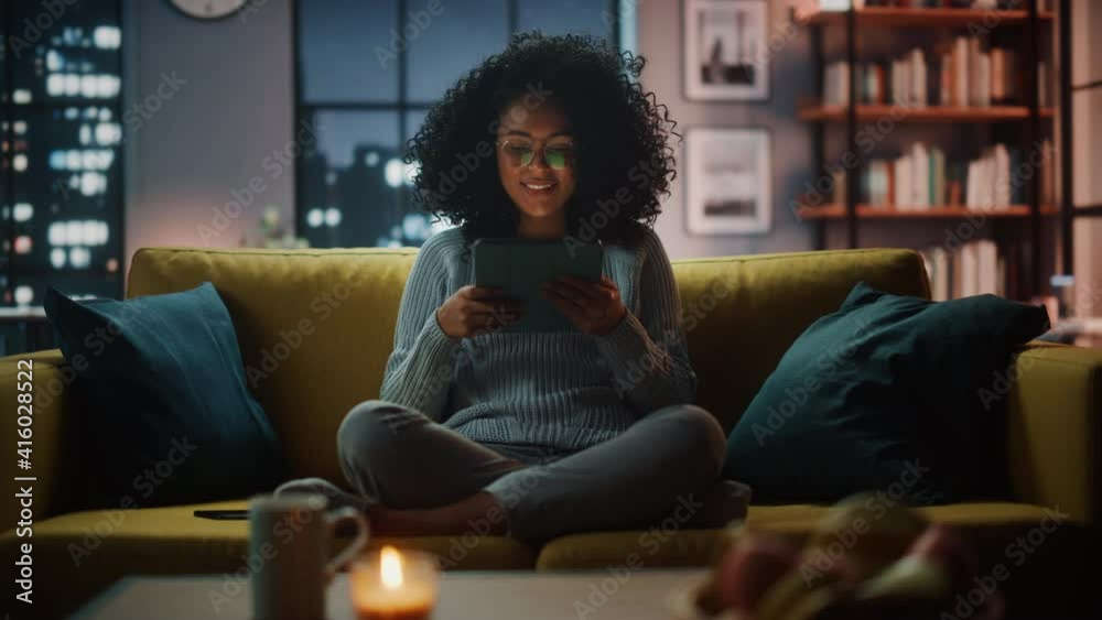 Beautiful Authentic Latina Female Sitting on Couch Sofa in a Cozy Dark Living Room in the Evening and Using Tablet Computer at Home. She's Browsing the Internet and Checking Videos on Social Networks. Beautiful Authentic Latina Female Sitting on Couch Sofa in a Cozy Dark Living Room in the Evening and Using Tablet Computer at Home. She's Browsing the Internet and Checking Videos on Social Networks.