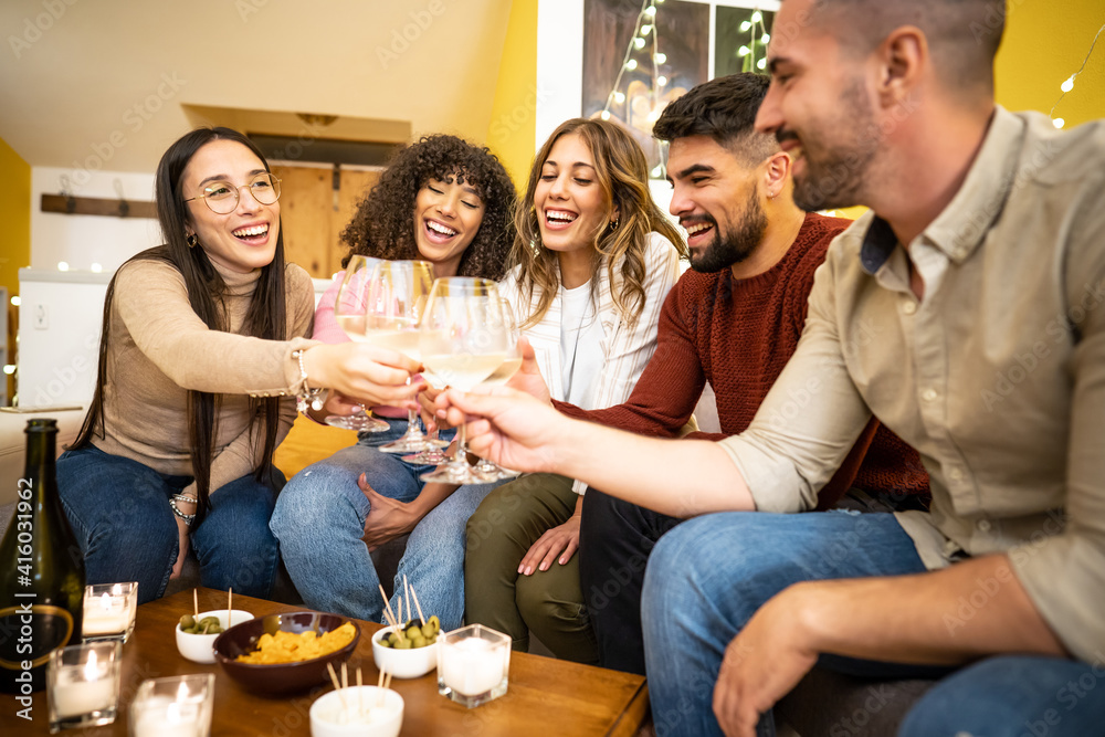 Multiracial young happy friends group at home sitting on the sofa toasting with champagne - Mixed race millennial laughing people celebrating clinking with wine glasses and table laden with snacks