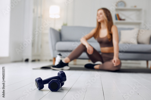 Blurred image of young beautiful athletic girl in leggings and top makes an exercises at home. Healthy lifestyle. In the foreground dumbbells.
