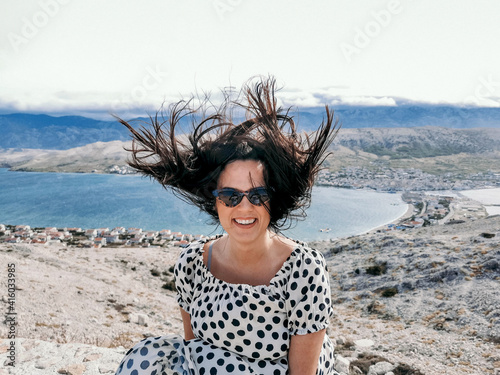 Smiling Caucasian woman in a polka dots flowy dress and sunglasses with hair blown up by the wind
