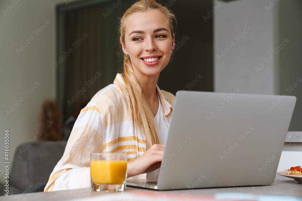 Smiling woman working with laptop while having breakfast at home Smiling woman working with laptop while having breakfast at home
