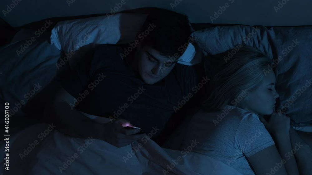 Cheating husband. Couple issue. Infidelity betrayal. Disloyal man using mobile phone for watching porn xxx film sex chat lying in bed with sleeping wife late at night in dark blue light.