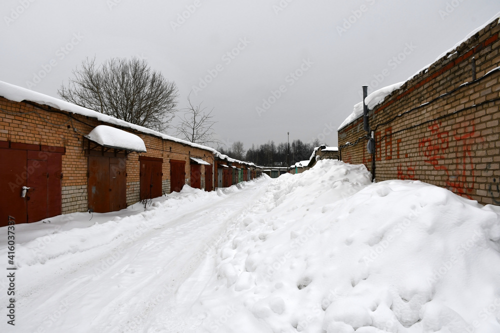 A cloudy winter day. Snowfall. Rows of one-story brick garages with closed metal painted gates. Drifts of snow near the walls along the road. Snow on the roofs.
