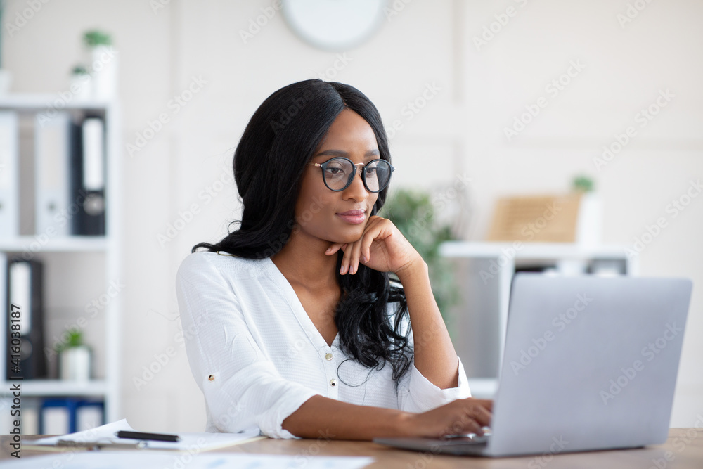 Positive female manager using laptop computer at workplace, typing document, taking part in online business meeting