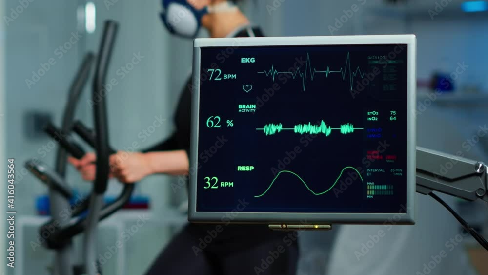 Monitor showing EKG scan of athlete running on cross trainer and medical specialists supervise exercise in background, controlling physical activity, measuring heart rate in sports science laboratory