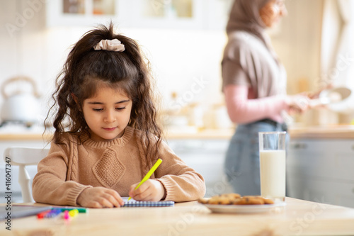 Adorable Little Girl Drawing In Kitchen While Her Muslim Mom Cooking Food