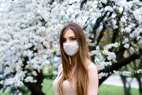 Close up portrait of tender girl in a white blouse under a blossoming cherry tree with a mask from the coronavirus.