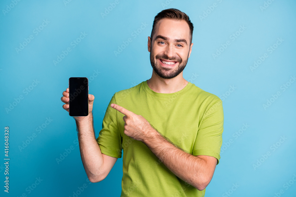 Photo of charming cheerful man point finger hold phone empty space show screen isolated on blue color background Photo of charming cheerful man point finger hold phone empty space show screen isolated on blue color background