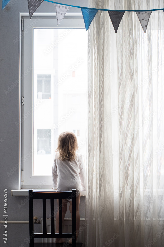 Lonely little girl at home stands at the window with her back to the camera. autism awareness day Lonely little girl at home stands at the window with her back to the camera. autism awareness day