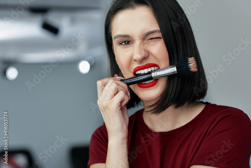Funny brunette female with red lips bites a brush making make-up. Happy young woman making herself makeup with a brush with organic cosmetics.