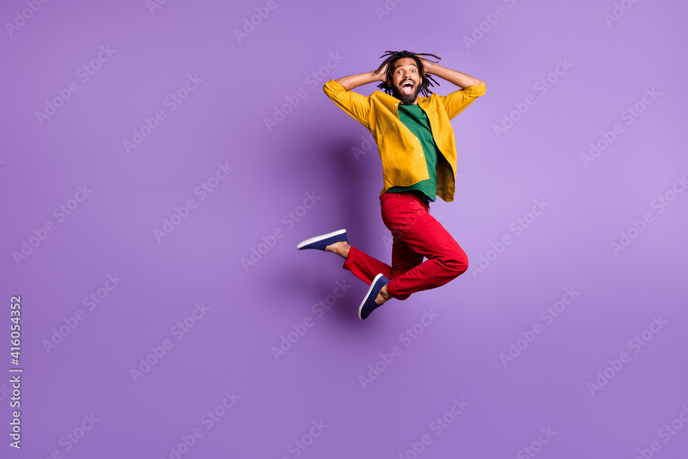 Full length photo of impressed dark skin man wear yellow shirt jumping high arms head isolated violet color background Full length photo of impressed dark skin man wear yellow shirt jumping high arms head isolated violet color background