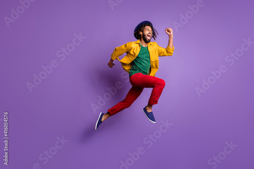 Full length photo of funky dark skin guy dressed yellow shirt headwear jumping running fast isolated purple color background