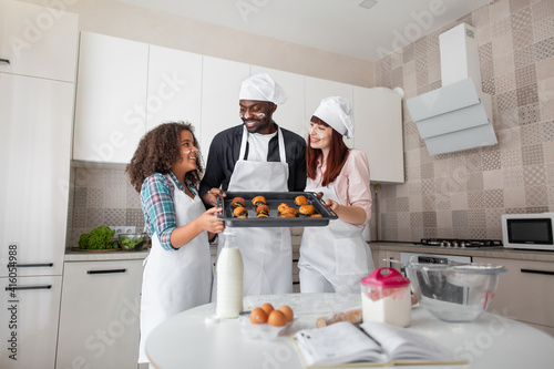 Cook at home and family bakery concept. Parents and daughter wearing aprons holding baking tray with fresh tasty pastry in kitchen at home