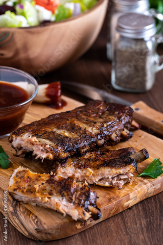 Close up of a rack of spareribs on a rustic wooden board with barbecue sauce and salad in behind.