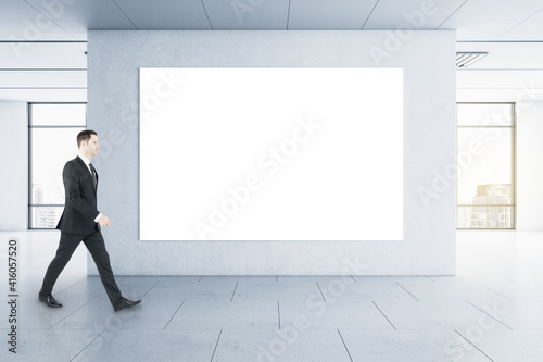 Businessman in black suit goes by blank white poster on the wall in the center of empty hall with city view