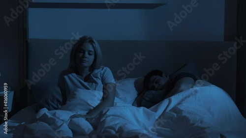 Night insomnia. Sleep disorder. Fatigue anxiety. Exhausted tired bored woman sitting in bed awake with peaceful resting man lying late in dark bedroom with blue light.
