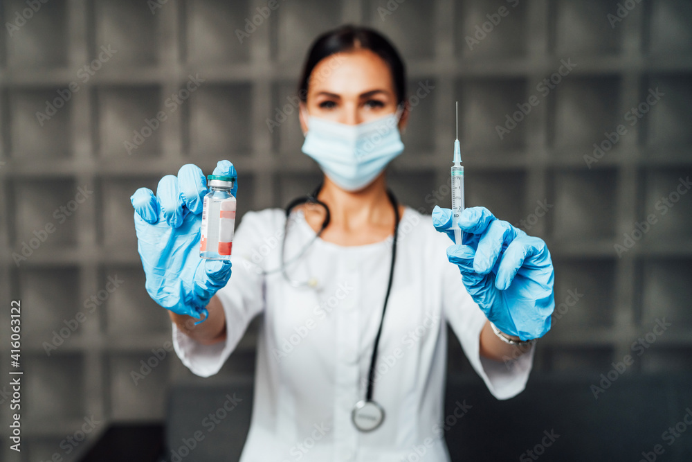 Naklejka premium The focus is on the nurse's hands in protective gloves holding the vaccine and the injection, in the background a nurse in a protective mask. Safe vaccination