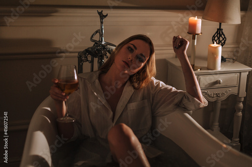 Young beautiful woman sitting in bathroom in expensive bathtub bath drinking wine looking at the camera