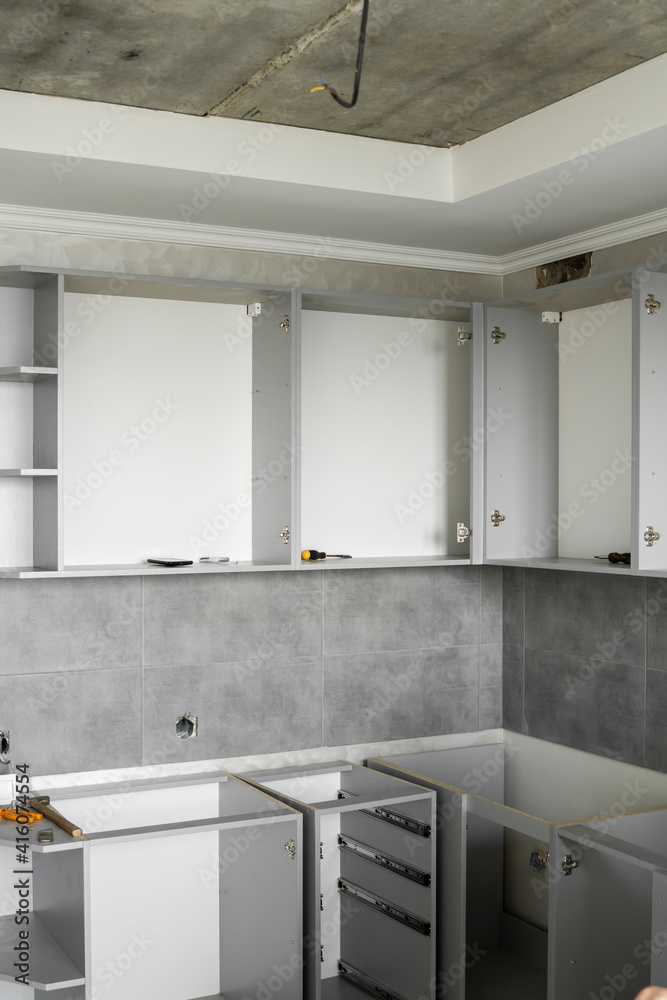 Custom kitchen cabinets installation without a furniture facades mdf. Gray modular kitchen from chipboard material on a various stages of installation in kitchen with a grey tile on floor and walls. Custom kitchen cabinets installation without a furniture facades mdf. Gray modular kitchen from chipboard material on a various stages of installation in kitchen with a grey tile on floor and walls.