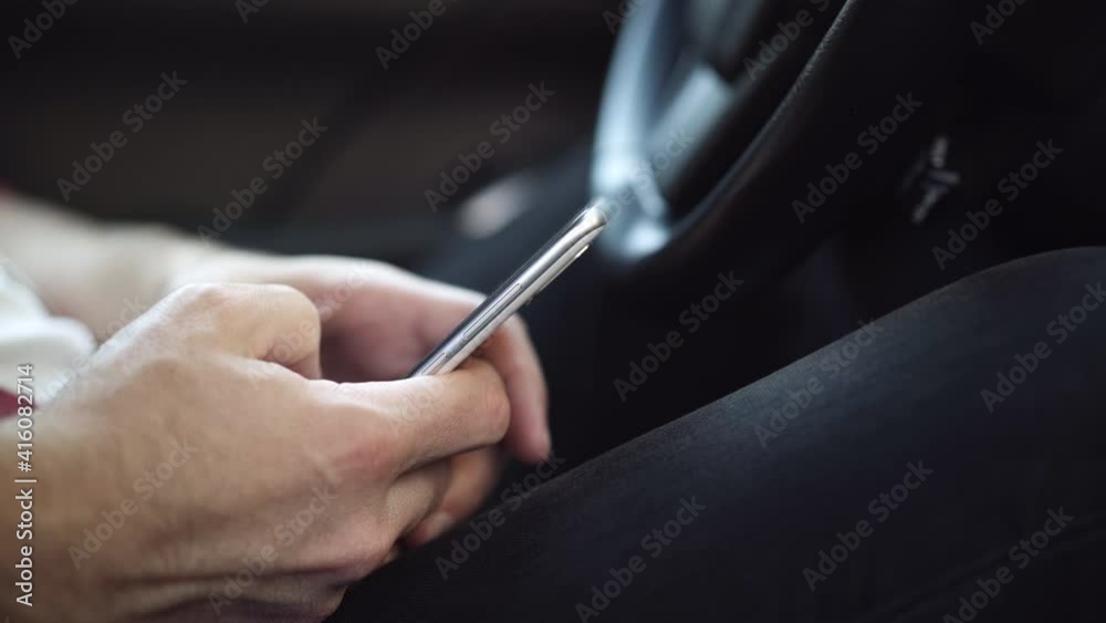 Close-up of male hands messaging online on smartphone with vehicle steering wheel at background. Unrecognizable Caucasian man typing message in social media sitting in car on driver's seat. Close-up of male hands messaging online on smartphone with vehicle steering wheel at background. Unrecognizable Caucasian man typing message in social media sitting in car on driver's seat.