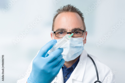 male doctor with face mask and medical gloves holding a syringe in front of a clinic room