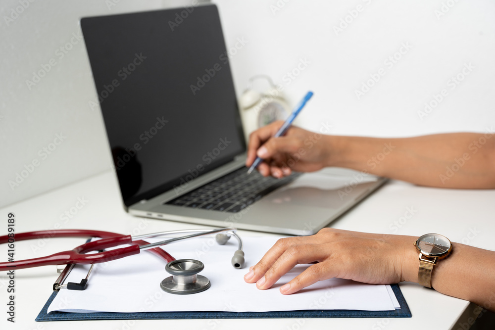 a doctor working on a laptop taking notes at the workplace. A female physician writing in a notebook using computer sitting at desk. a doctor working on a laptop taking notes at the workplace. A female physician writing in a notebook using computer sitting at desk.
