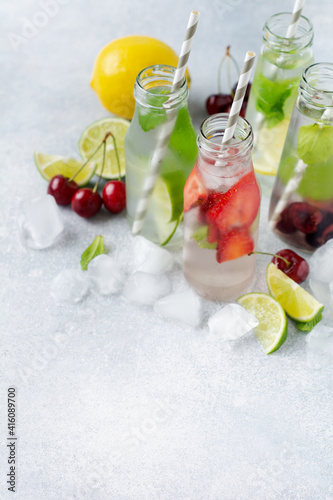 Lots of bottles with refreshing summer lemonade with lime, strawberry, cherry, cucumber and ice on a gray concrete background