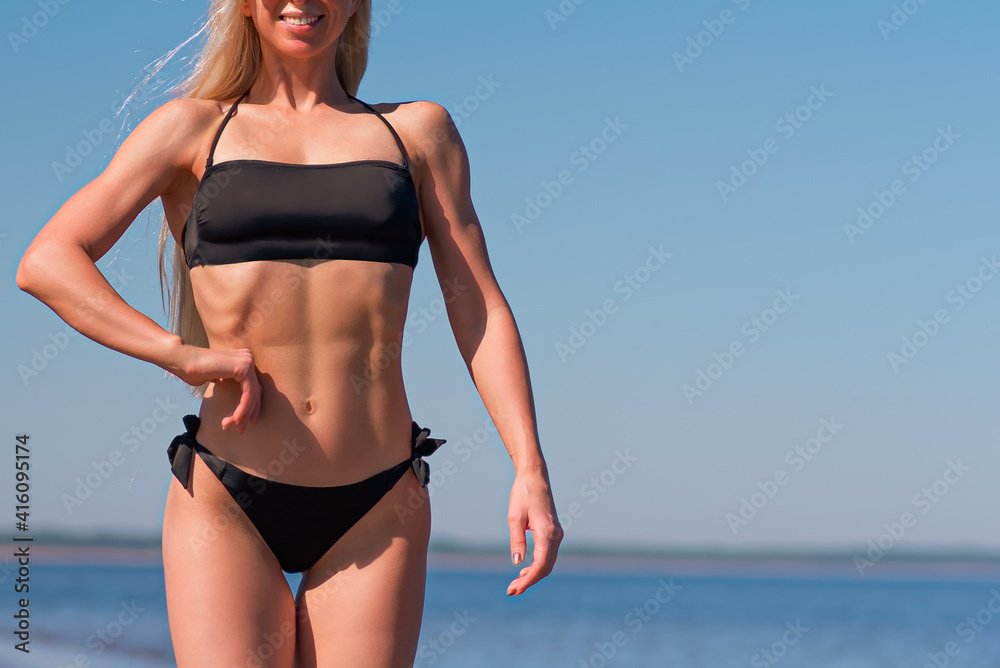 sporty woman in a black swimsuit walks along the beach against the background of blue sky and sea. sport and recreation concept