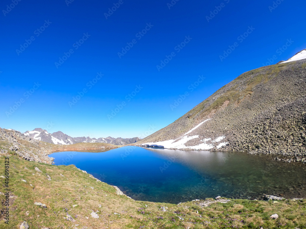 Clear, navy blue lake hiding between tall mountain peaks. Some of the slopes are still covered with snow. In the back here is another mountain range. Clear and bright day. Perfect condition for hiking